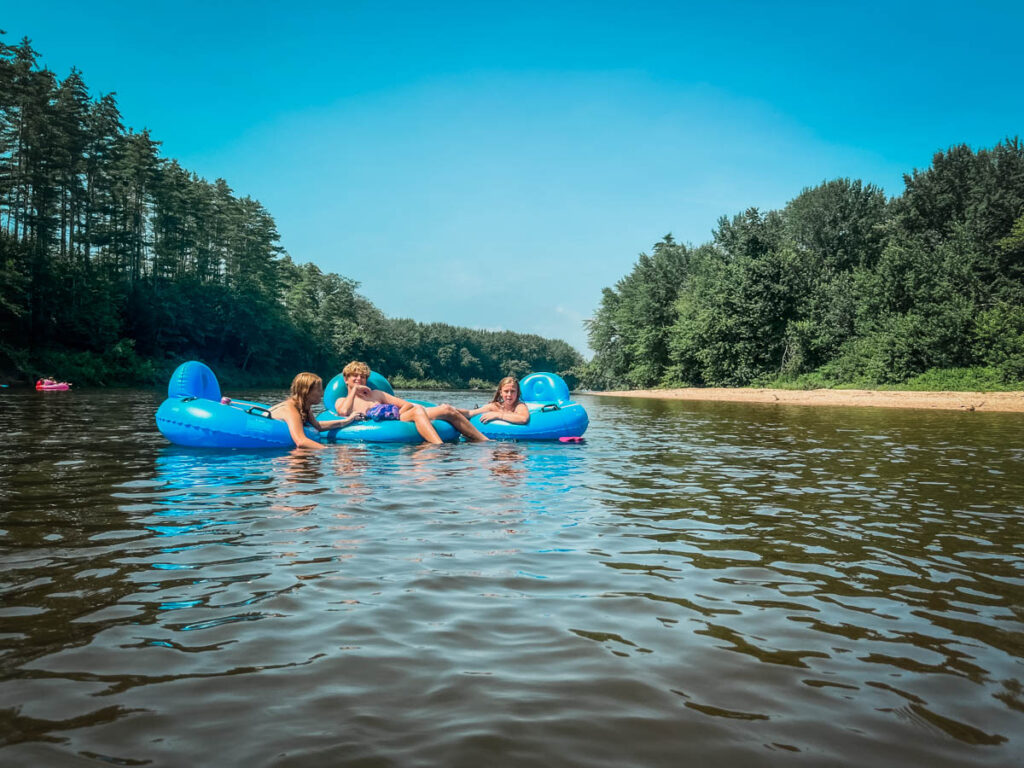 Three people relaxing in bright blue inflatable tubes on the Saco River near North Conway, New Hampshire, with forested riverbanks.