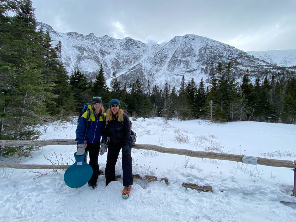 Two winter hikers standing in the snow with the snow-covered mountains of Tuckerman Ravine on Mount Washington behind them.